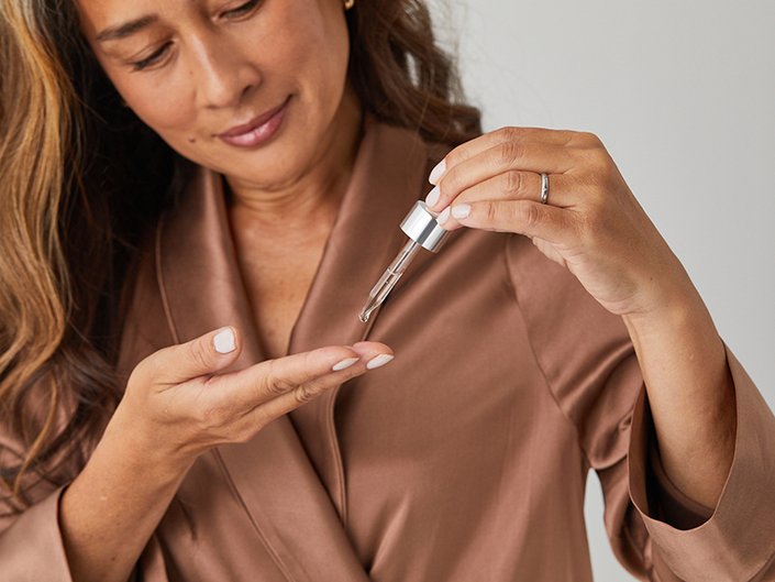 Person with long dark hair wearing a brown satin robe and dropping serum onto their fingers with a dropper