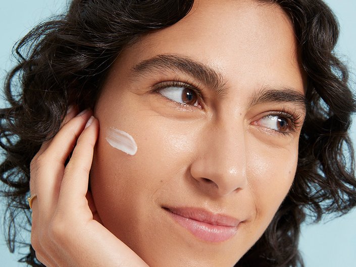 Person with dark curly hair with face cream swatched on their cheek