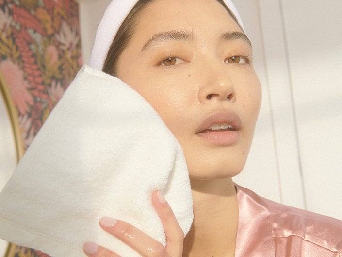 Person wearing a white headband and pink satin robe blotting their cheek with a white washcloth
