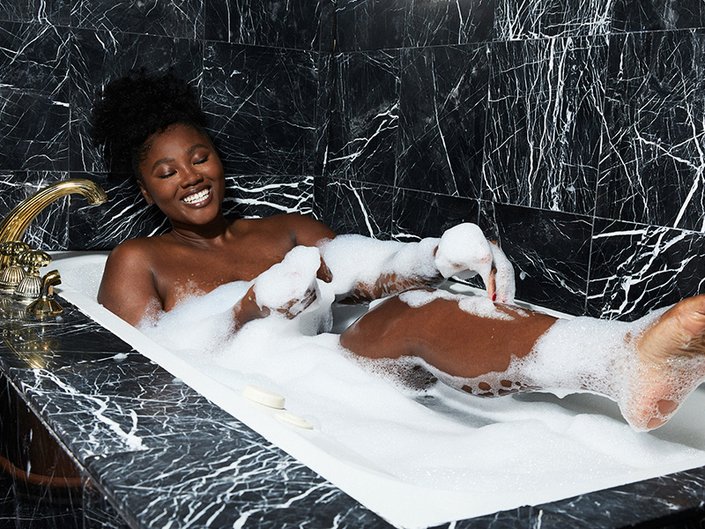 Person sitting in a bubble bath and smiling with one leg out of the water, touching their leg