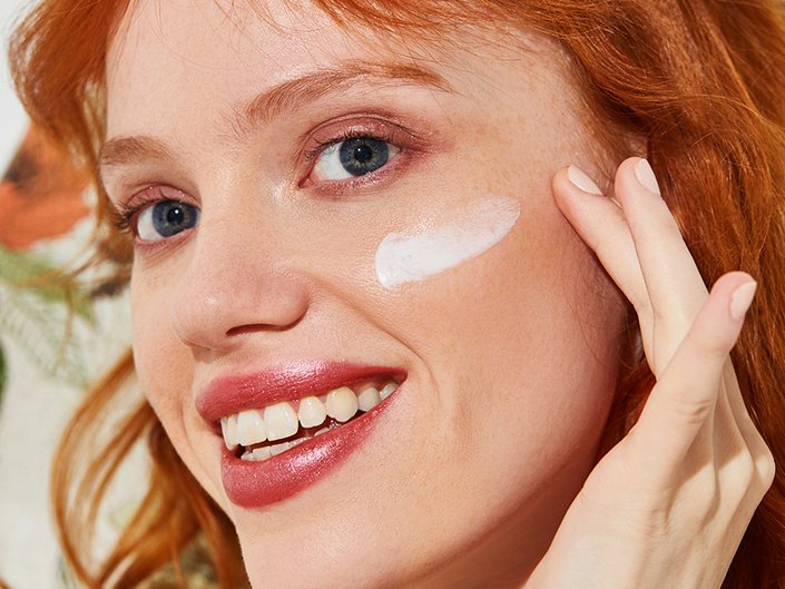 Model with red hair and blue eyes applying a swatch of SPF to face.